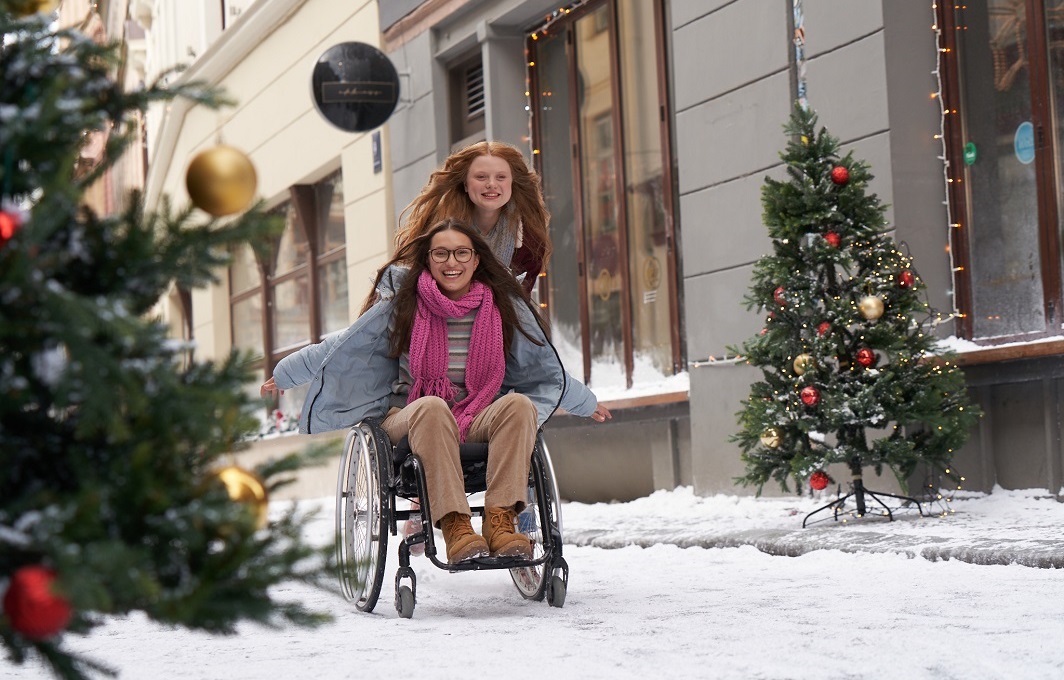 Ein Mädchen schiebt ein anderes Mädchen im Rollstuhl durch Schnee und eine Straße mit Weihnachtsschmuck. Beide sind fröhlich.