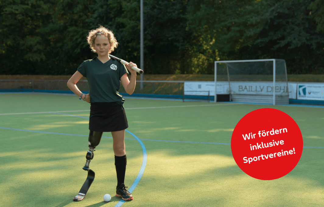 Ein junges Mädchen mit Beinprothese steht mit ihrem Hockeyschläger auf dem Hockeyplatz.