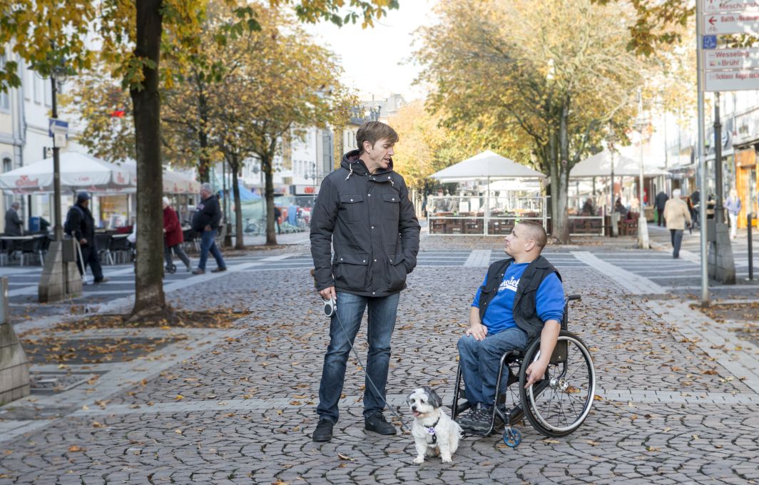 Zwei Männer stehen auf einem Platz in der Kleinstadt. Einer von den Männern sitzt im Rollstuhl.