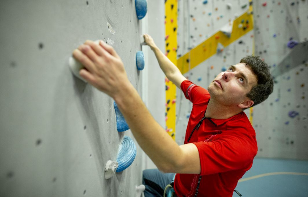 Ein Mann klettert eine Kletterwand hoch. Er hält sich an bunten Griffen fest.