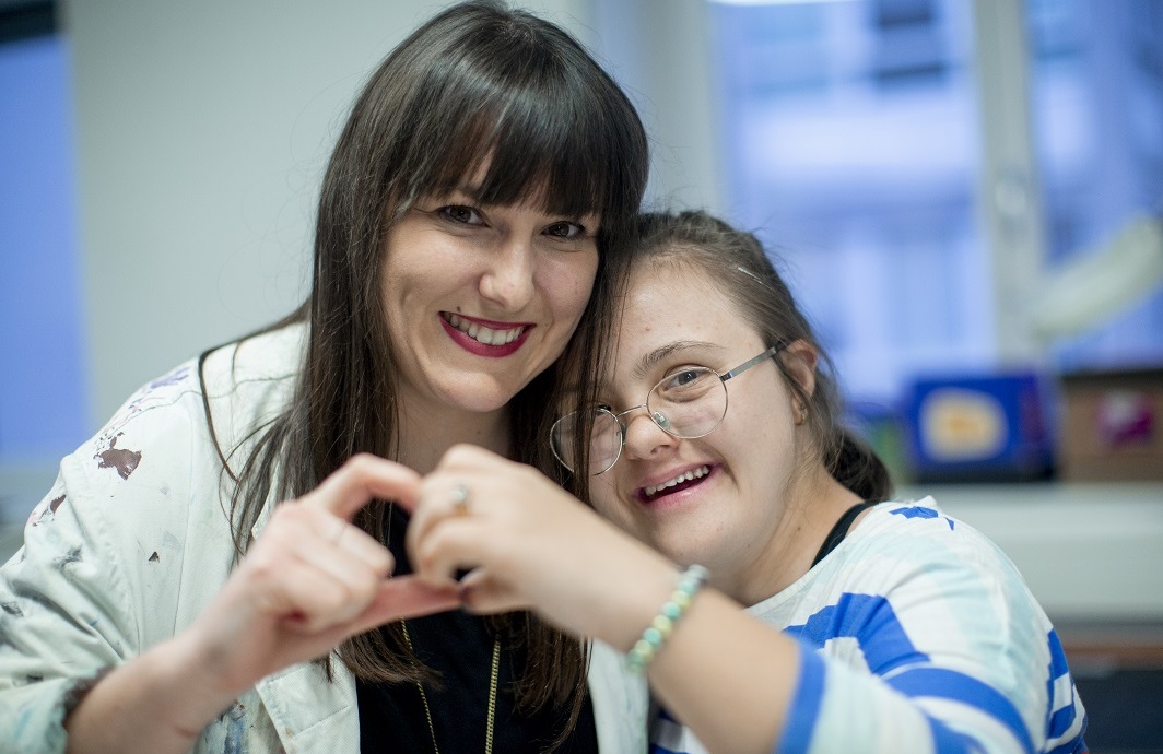 Ein Kind mit Downsyndrom und ihre Betreuerin formen mit ihren Händen ein Herz