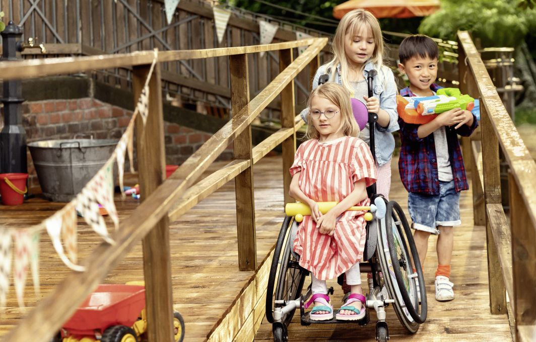 Drei Kinder spielen auf einem Spielplatz. Ein Maedchen sitzt im Rollstuhl.
