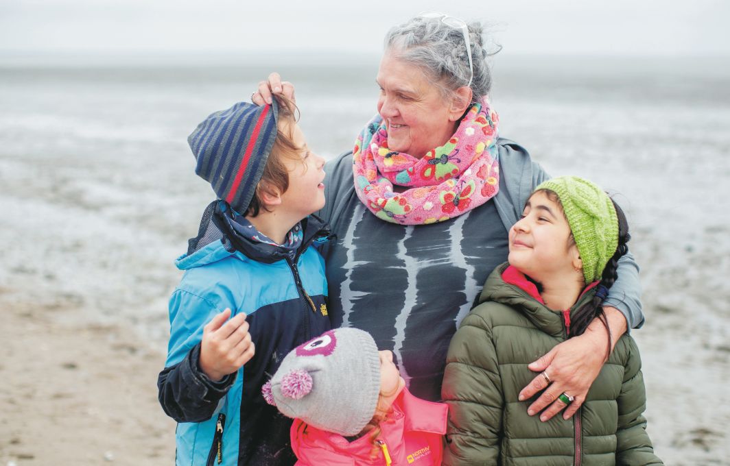 Eine Frau und drei Kinder stehen am Strand. Die Frau hat die Arme und die Kinder gelegt.