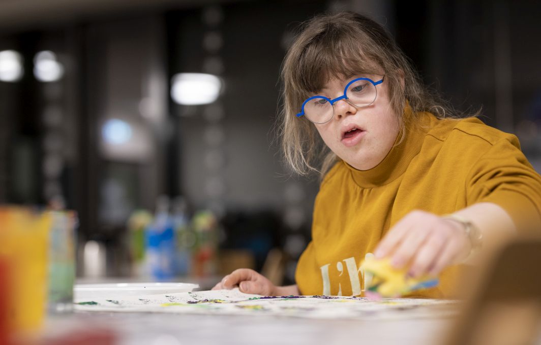 Eine Frau mit Downsyndrom udn Brille malt konzentriet auf einem Blatt Papier.