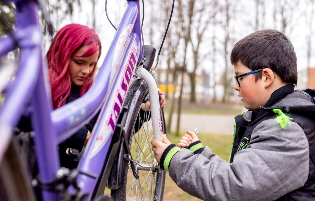 Ein Junge arbeitet mit einem Werkzeug an einem Fahrrad. Ein Mädchen im Hintergrund schaut zu.