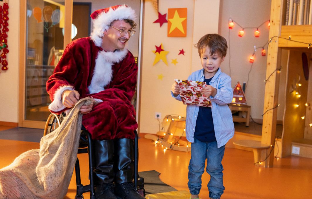 Ein Mann im Rollstuhl ist als Nikolaus verkleidet und überreicht einem Kind ein Geschenk.