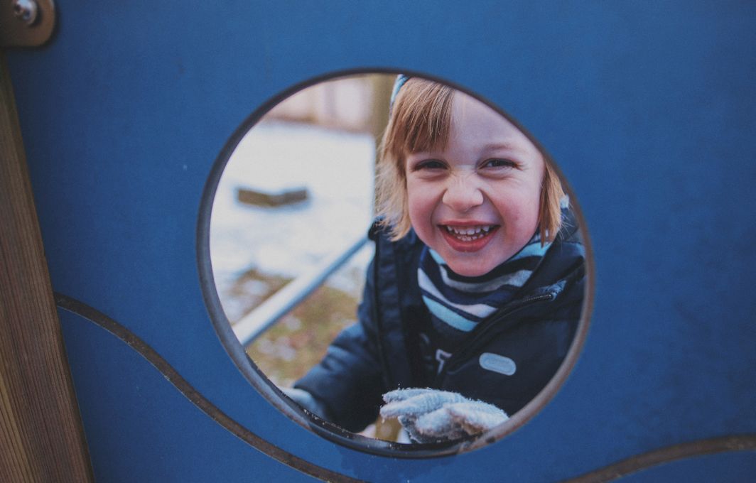 Ein Kind schaut durch ein Guck-Loch in einer Wand am Spielplatz und grinst dabei in die Kamera.
