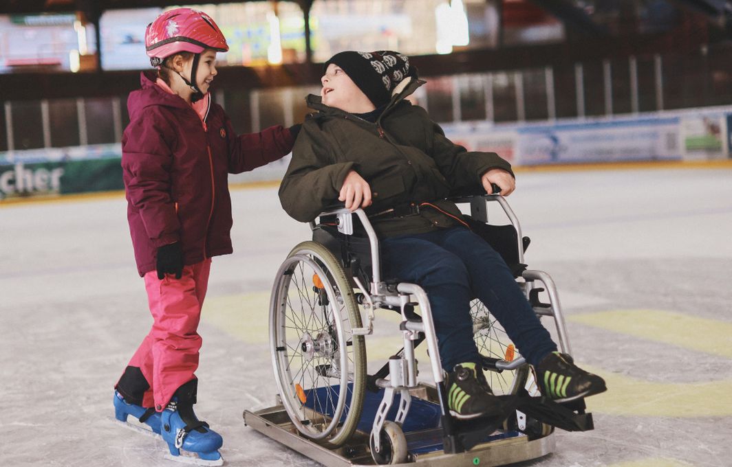 Zwei Kinder fahren über eine Eisfläche. Ein Kind sitzt im Rollstuhl, der auf einem Eisgleiter befestigt ist.