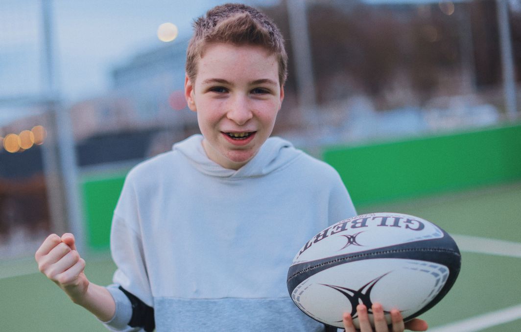 Ein Junge steht auf einem Sportplatz und hält einen Rugby in der Hand. Er lacht in die Kamera.