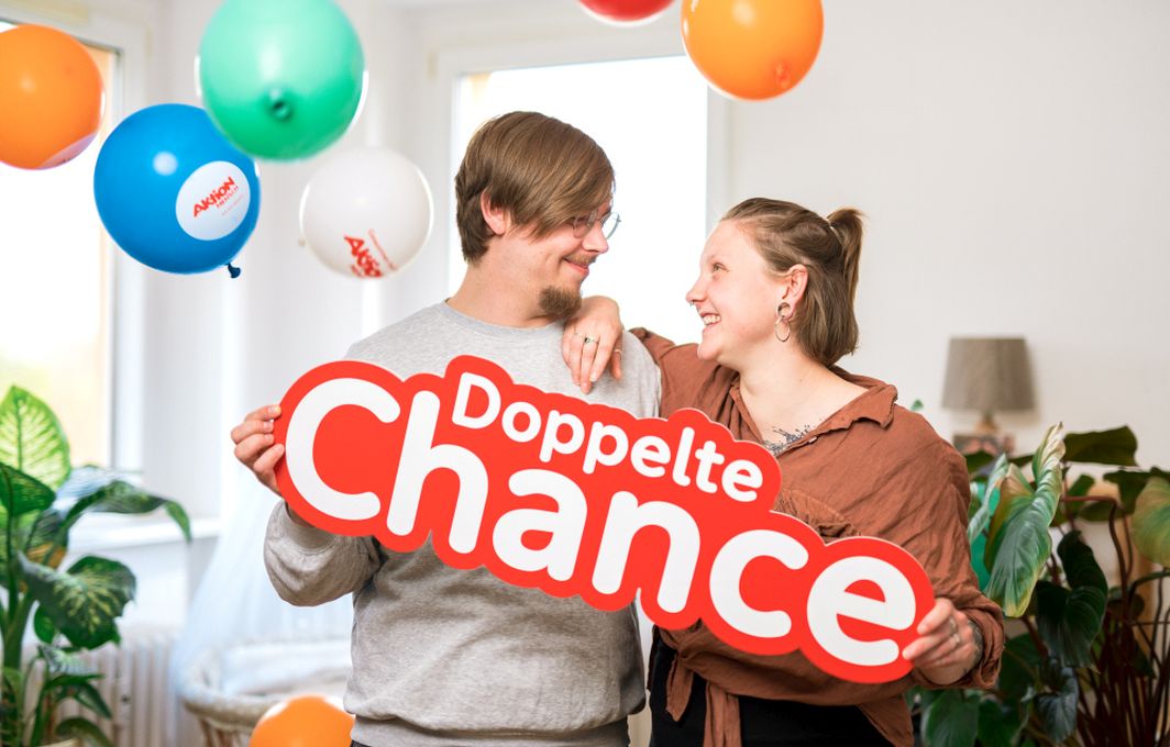 Gewinner*in Sandro und Fine stehen nebeneinander und lachen sich an. Sie halten ein Schild mit "Doppelte Chancen". Im Hintergrund sind bunte Luftballons.