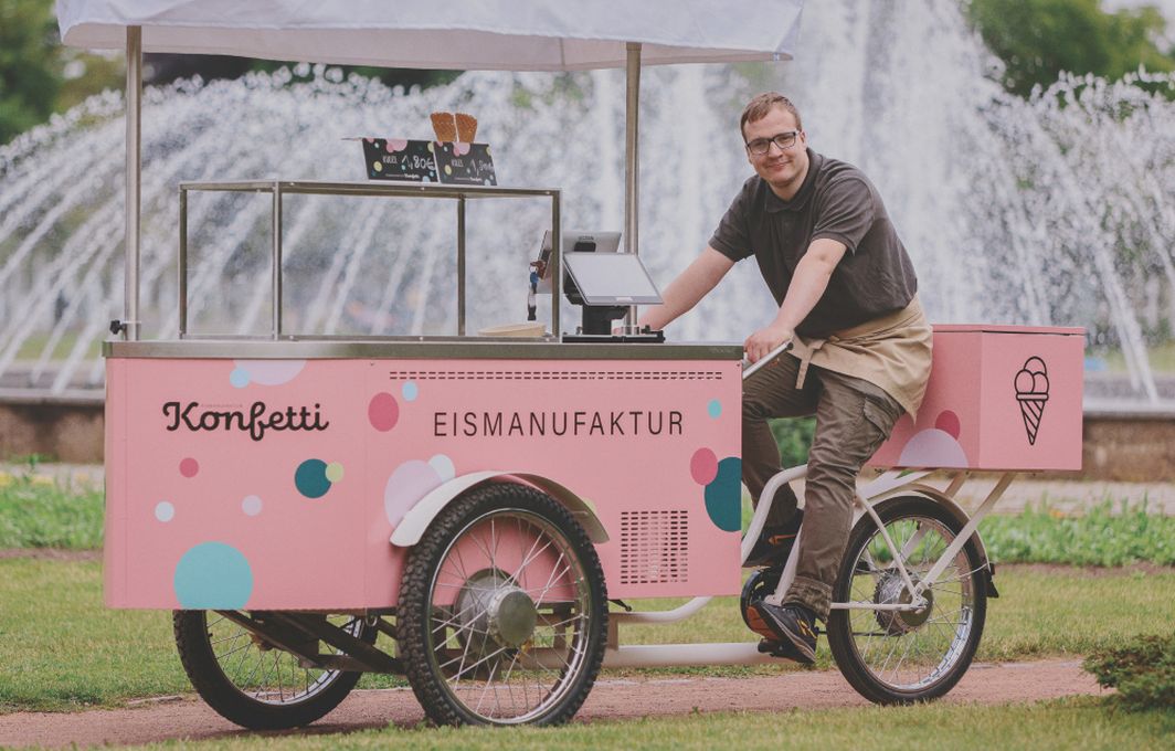 Ein Mann sitzt auf einem Lastenfahrrad. Vorne ist eine große Truhe für Eis montiert, darauf der Schriftzug "Konfetti Eismanufaktur." Im Hintergrund sieht man eine Parkanlage mit Springbrunnen.