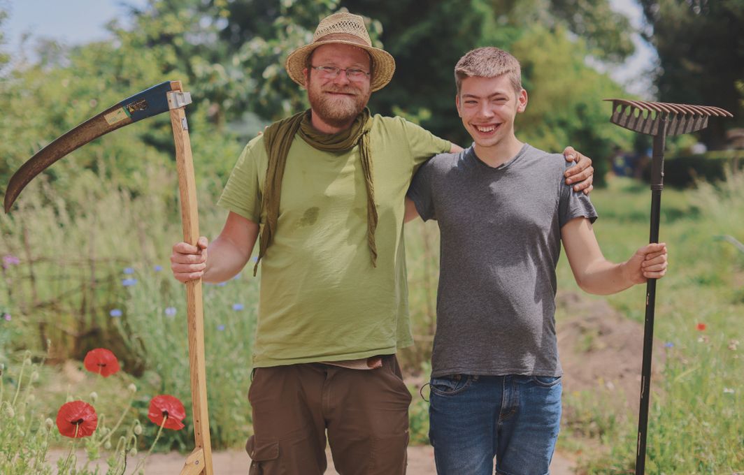 Der Gärtner Johannes und der Jugendliche Luis stehen Arm in Arm im Garten. Sie halten jeweils ein Arbeitsgerät in der freien Hand. Johannes trägt einen Hut. Beide lachen.