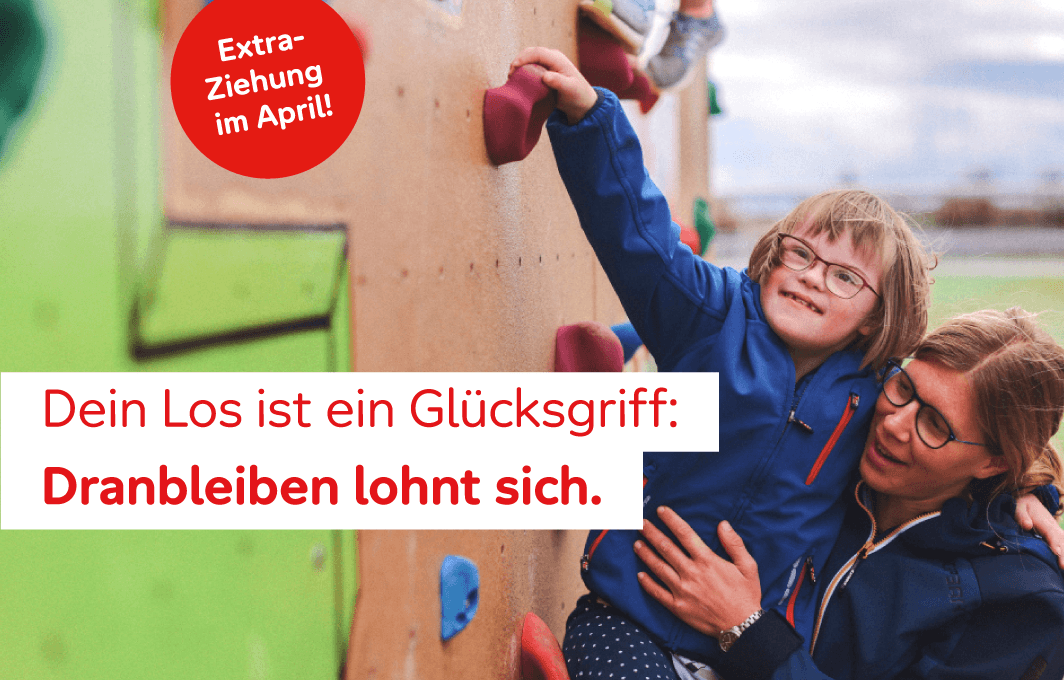 Eine Mutter hält ihre Tochter auf ihrem rechten Arm. Die Tochter greift mit ihrer Hand nach einem Stein in der Kletterwand und hat den anderen um den Hals ihrer Mutter gelegt.