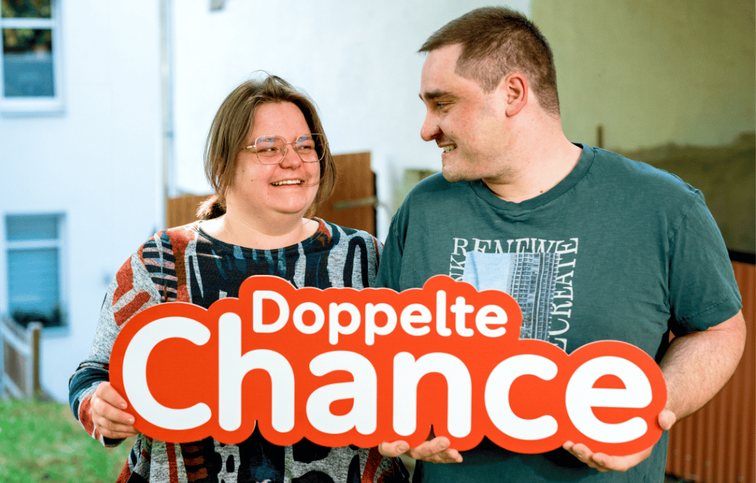 Eine Frau und ein Mann halten ein Schild in der Hand, auf dem "Doppelte Chance" steht.