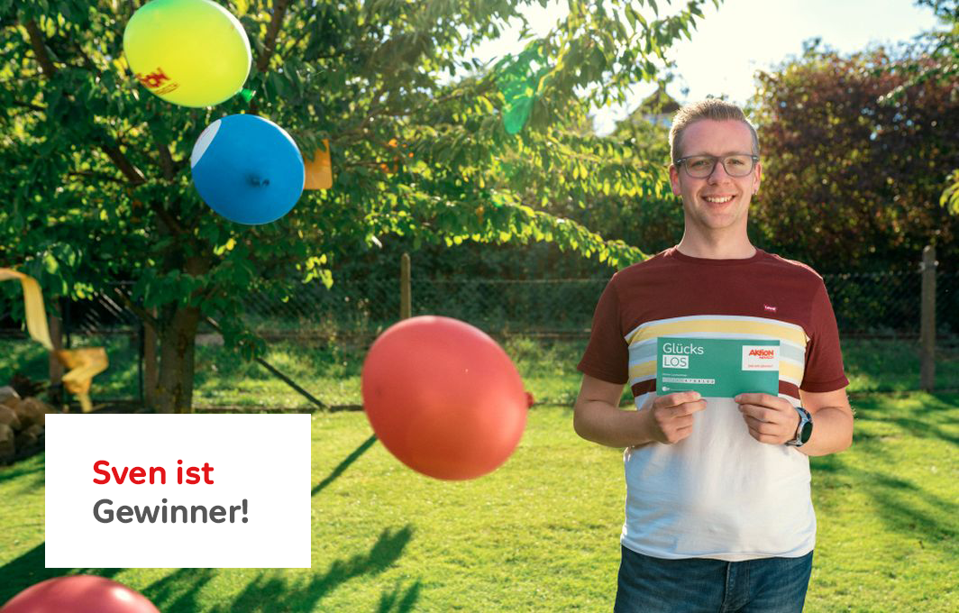 Gewinner Sven hält sein Glücks-Los in die Kamera und lacht. Um ihn herum fliegen bunte Luftballons.