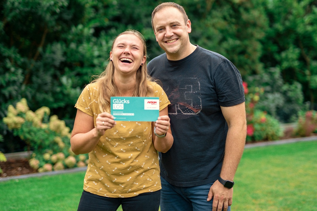 Gewinnerin Bozena und ihr Mann halten ein Glücks-Los in der Hand und strahlen in die Kamera.