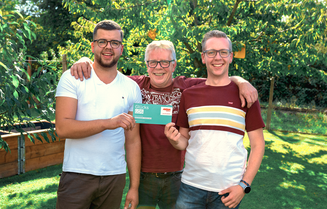 Drei Männer stehen im Garten und halten ein Glücks-Los in der Hand.