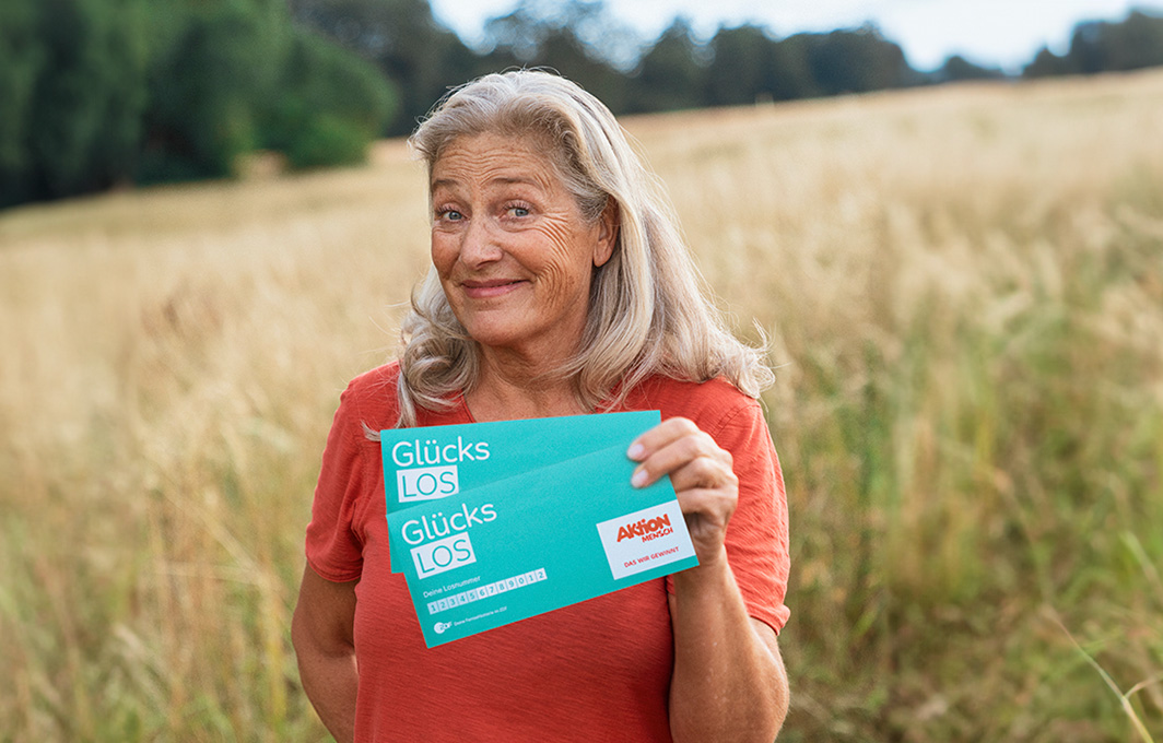 Eine Frau steht auf einem Feld und hält zwei Glücks-Lose in der Hand.