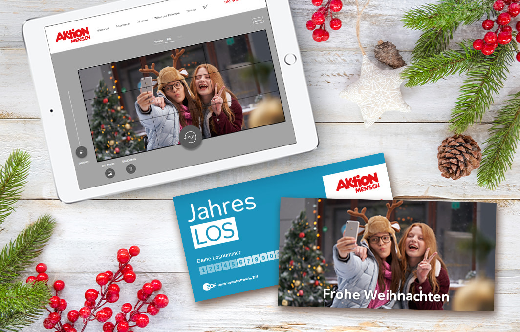 Es liegt ein Tablet auf dem Tisch, worauf die Benutzeroberfläche des Losdesigners abgebildet ist. Neben dem Tablet liegen zwei Jahreslose, eins mit dem im Losdesigner erstellten Motiv von zwei Mädchen, die ein Selfie machen.