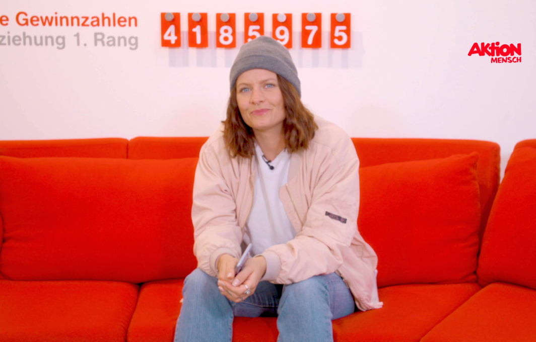 Mandy sitzt auf der Couch bei Aktion Mensch und hält ein Glück-Los in der Hand.
