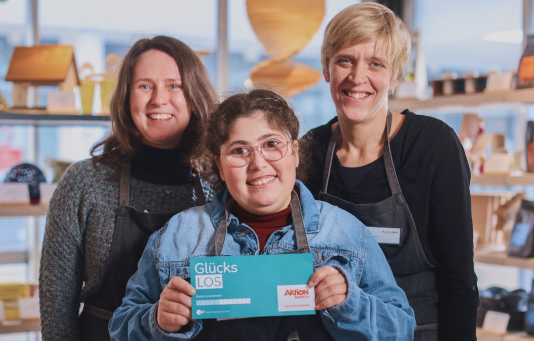 Drei Frauen stehen vor einem Marktstand und lachen in die Kamera. Die Frau in der Mitte hält ein Glücks-Los in der Hand.