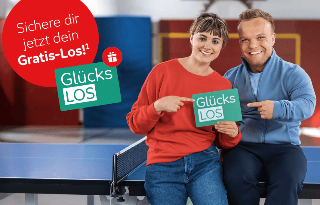 Niko Kappel und eine Dame lehnen an einer Tischtennisplatte und präsentieren das Glücks-Los. Der Link führt zur URL https://www.aktion-mensch.de/lotterie/gewinn