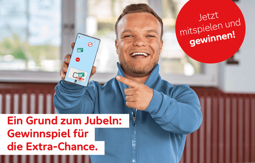 Niko Kappel zeigt grinsend auf seinen Handy-Bildschirm, auf dem ein Online-Spiel der Aktion Mensch zu sehen ist.
