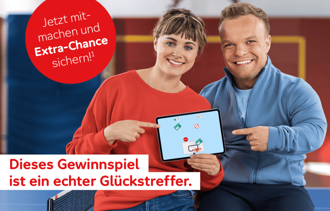 Niko Kappel und eine blonde Frau sitzen auf einer Tischtennisplatte und zeigen auf ein Tablet, auf dem das Aktion Mensch Korbleger-Spiel läuft.