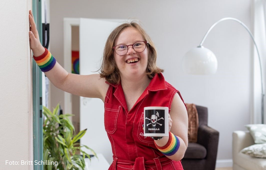 Natalie Dedreux steht lässig am Türrahmen und hält eine Tasse in der Hand. Sie lacht in die Kamera.