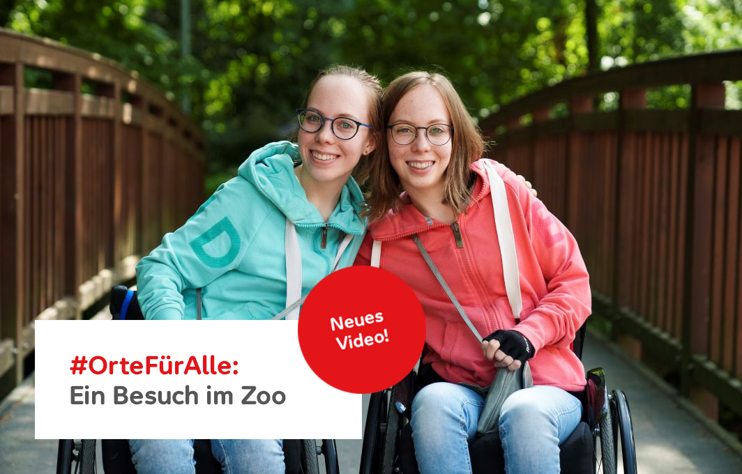 Leona und Ellen lachen in die Kamera. Beide sitzen im Rollstuhl. Im Hintergrund sieht man grüne Hecken.