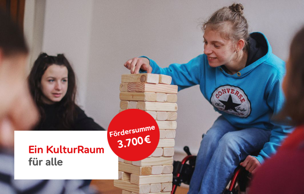 Ein Mädchen sitzt im Rollstuhl und spiel mit einem Jenga-Turm, der vor ihr auf dem Boden steht. Im Kreis drum herum sitzen andere Mitspieler*innen.