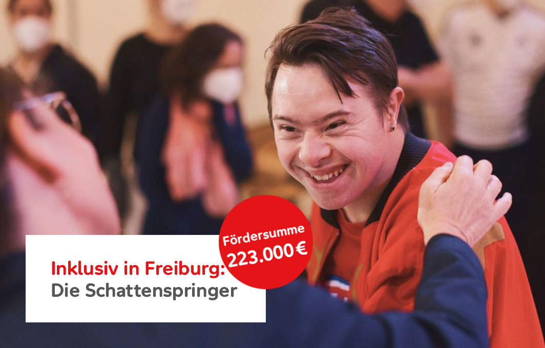 Eine Person mit Down-Syndrom lacht eine andere Person an, die ihr den Arm auf die Schulter legt.