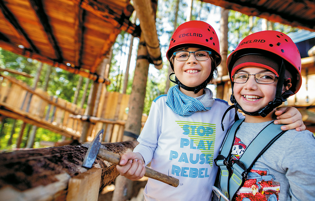 Zwei Kinder im Baumhaus. Eins von ihnen hält einen Hammer in der Hand. Beide tragen einen roten Helm.
