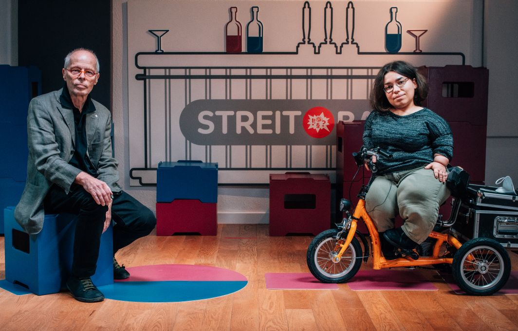 In einem Aufnahmestudio sitzen ein älterer Mann mit Halbglatze und Brille und eine junge Frau mit mittellangem, dunklen Haar. Die Frau sitzt auf einem Fahrrad mit drei Rädern. Hinter ihnen an der Wand steht der Schriftzug "Streitbar".