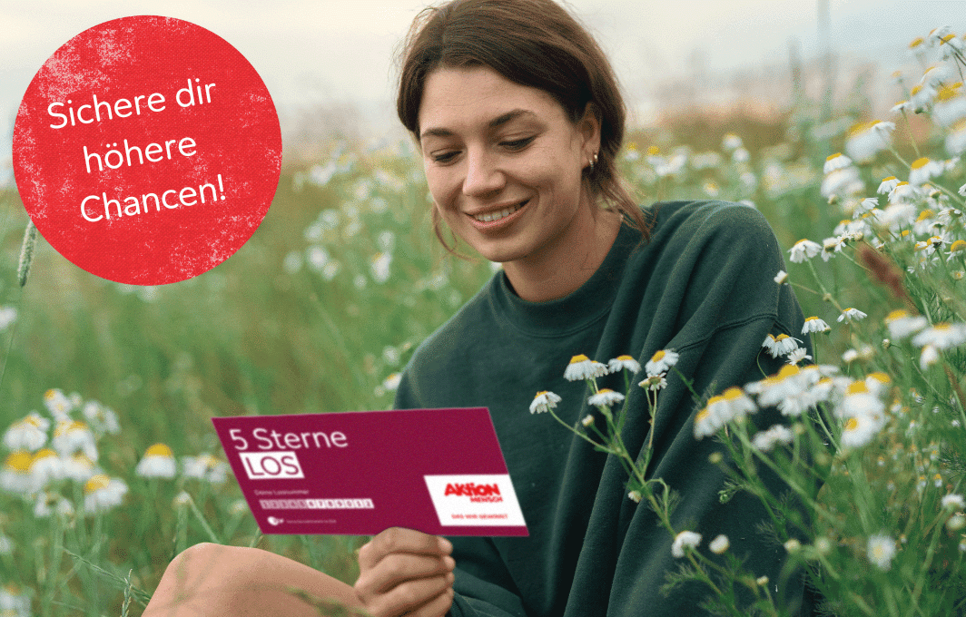 Eine junge Frau sitzt auf einer grünen Wiese und hält ein 5 Sterne-Los in der Hand.