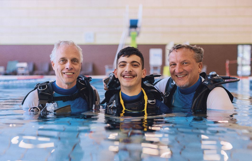 Links ist Peter, in der Mitte ein Tauchschüler und rechts der Tauchlehrer. Ihre Köpfe schauen aus dem Wasser des Schwimmbeckens. Sie tragen Neoprenanzüge