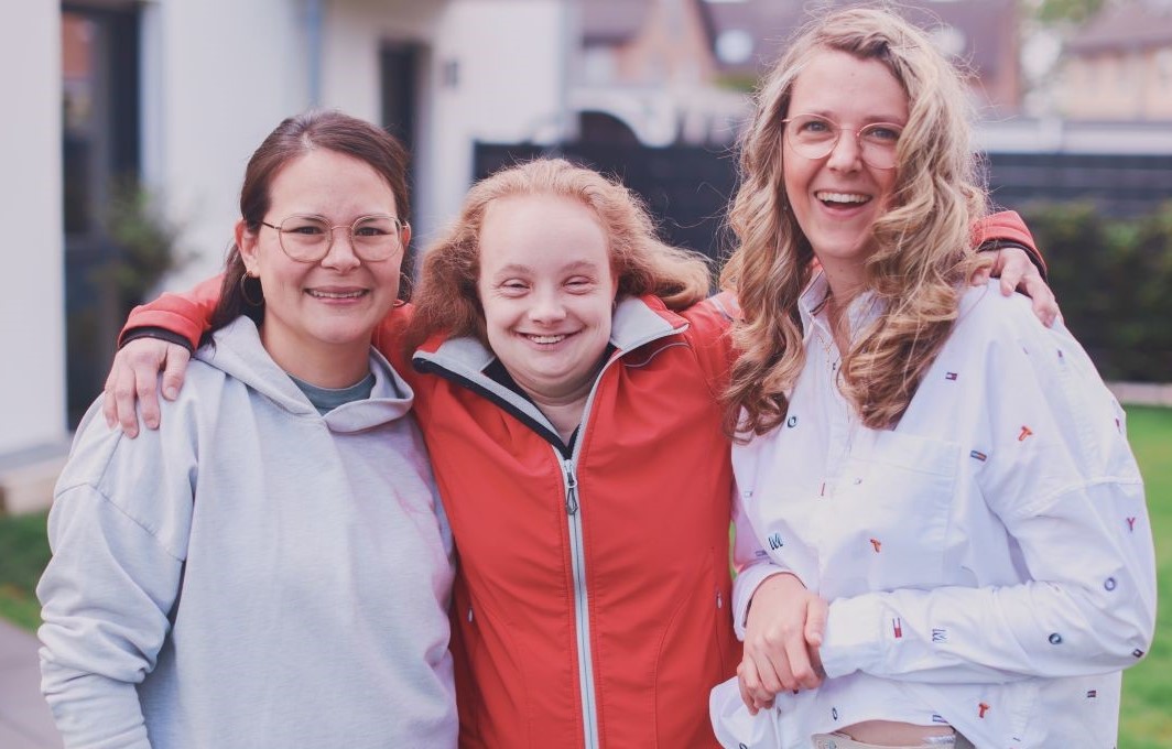 Drei Frauen stehen fröhlich lächelnd beieinander, die mittlere hat ihre Arme auf die Schultern der anderen beiden gelegt.
