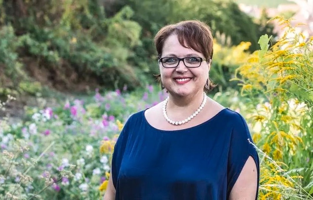 Eine Frau mit blauem Oberteil und weißer Perlenkette steht vor bunten Blumen im Garten