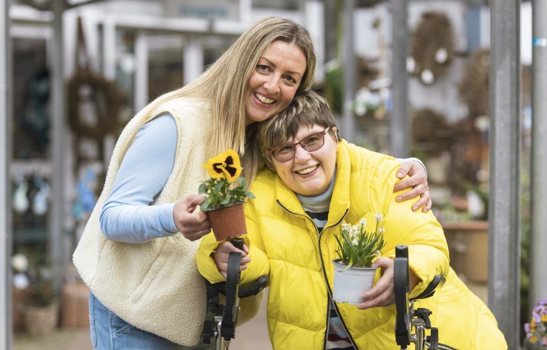 Zwei Frauen stehen Arm in Arm nebeneinander und halten Blümchen in die Kamera.