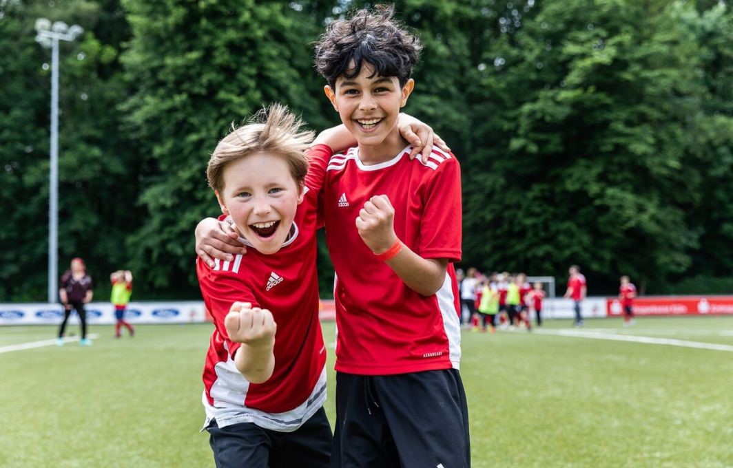 Zwei Jungs im Fußballtrikot stehen Arm in Arm nebeneinander auf dem Platz und jubeln in die Kamera. Hinter ihnen sieht man verschwommen weitere Teammitglieder