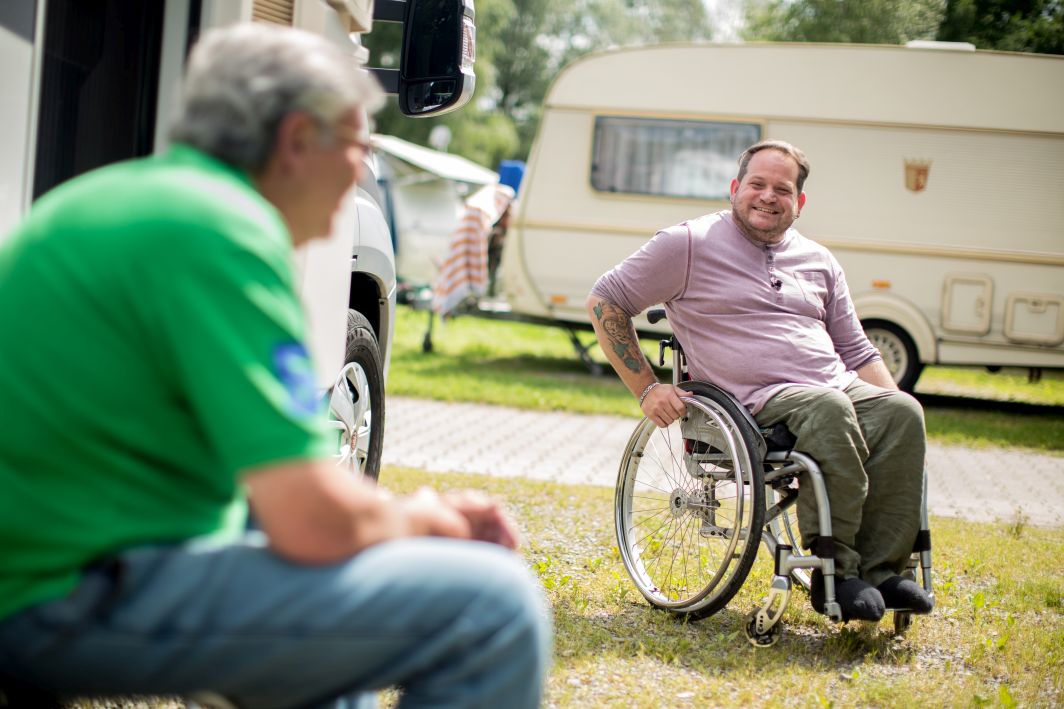 Ein Mann mit Tattoos und Rollstuhl lacht auf dem Campingplatz einen Urlauber an, der vor seinem Wohnmobil sitzt.