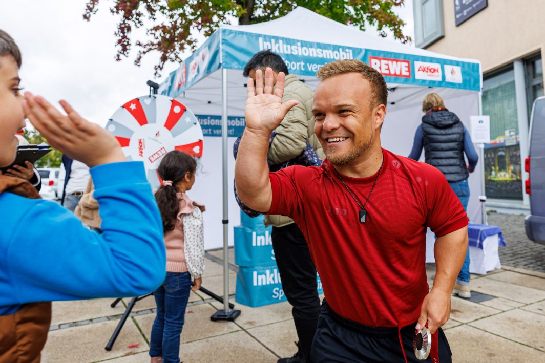Niko Kappel, der erfolgreiche Parasportler im Kugelstoßen, gibt einem Jungen fröhlich ein High Five. Hinter ihm ist der Aufbau des Inklusionsmobils zu sehen.