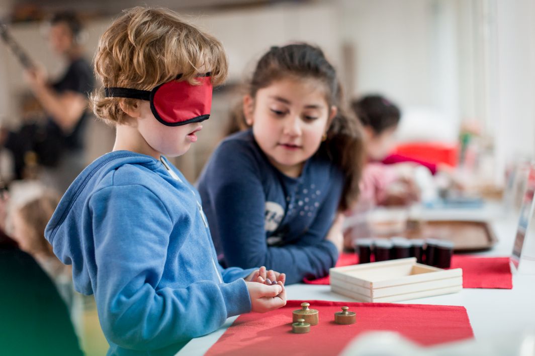 Verlinkte Aufnahme aus der Lernwerkstatt: Ein Kind mit Schlafmaske wiegt in seinen Händen unterschiedlich schwere Gegenstände ab. Ein Mädchen schaut ihm dabei zu. Der Link führt zur Seite des Förderprojekts.