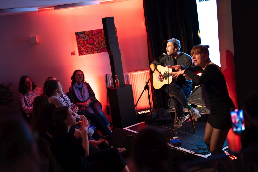 Max Giesinger performt mit seiner Gitarre auf einer kleinen Bühne vor kleinem Publikum. Neben ihm steht eine Deaf-Performerin. Der Link führt zu einem Artikel über das Privatkonzert in Köln.