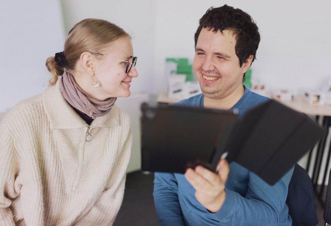 Eine junger Mann mit Behinderung hält lächelnd ein Tablet hoch, neben ihm sitzt eine junge Frau, die ihn anlächelt. Der Link führt zur Seite über "Digitale Teilhabe an Gesundheitsbildung".