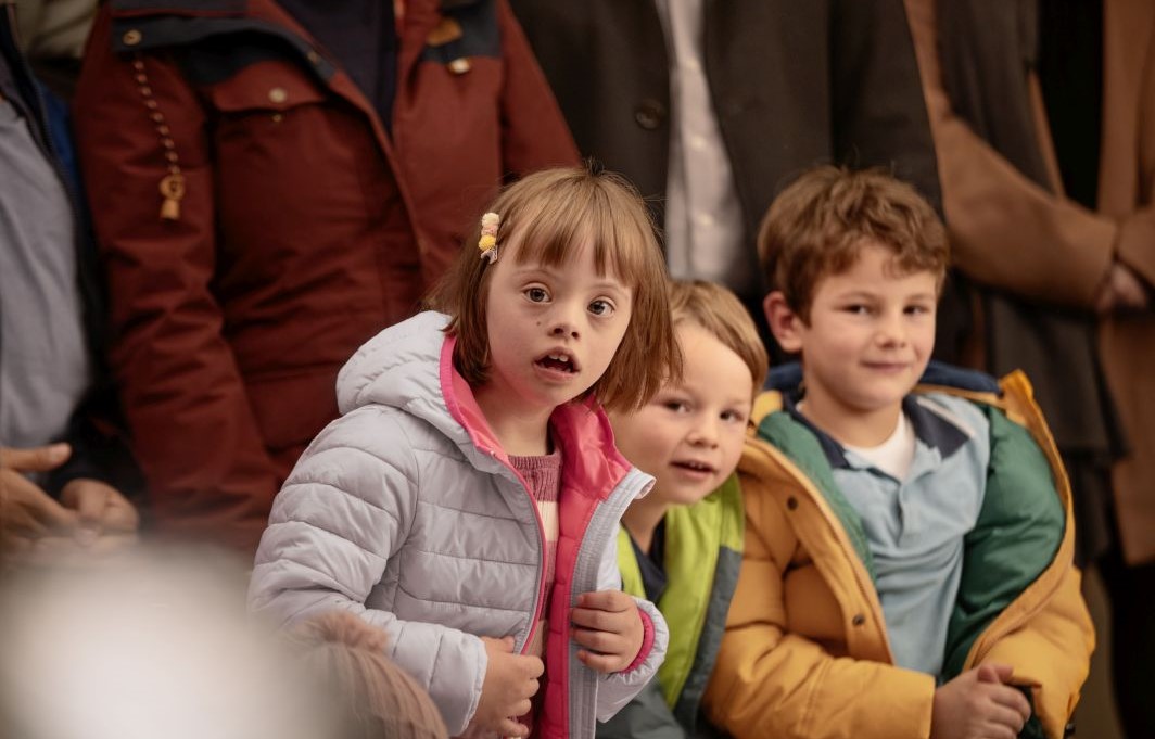Drei Kinder, eines von ihnen mit Down-Syndrom, schauen voller Spannung in dieselbe Richtung. Hinter ihnen stehen einige Erwachsene.
