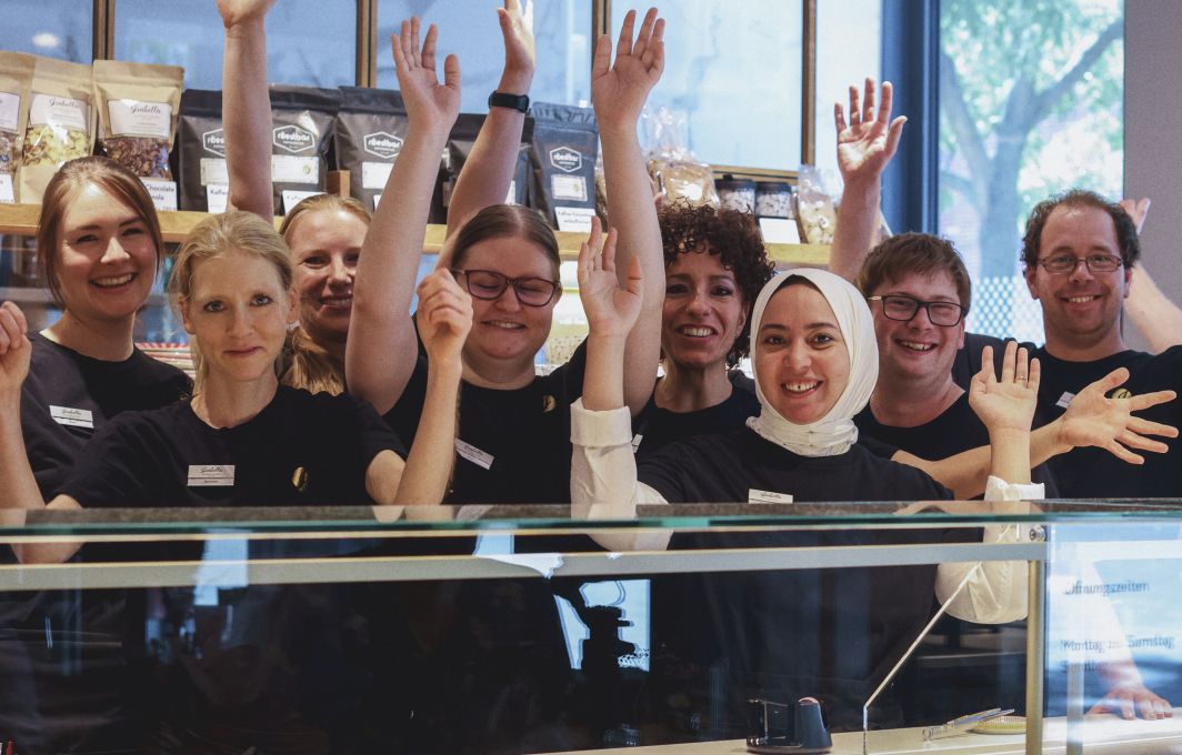 Eine Gruppe von Menschen unterschiedlichen Geschlechts und Hautfarbe und mit und ohne Behinderung stehen lachend hinter einer Patisserie-Theke.