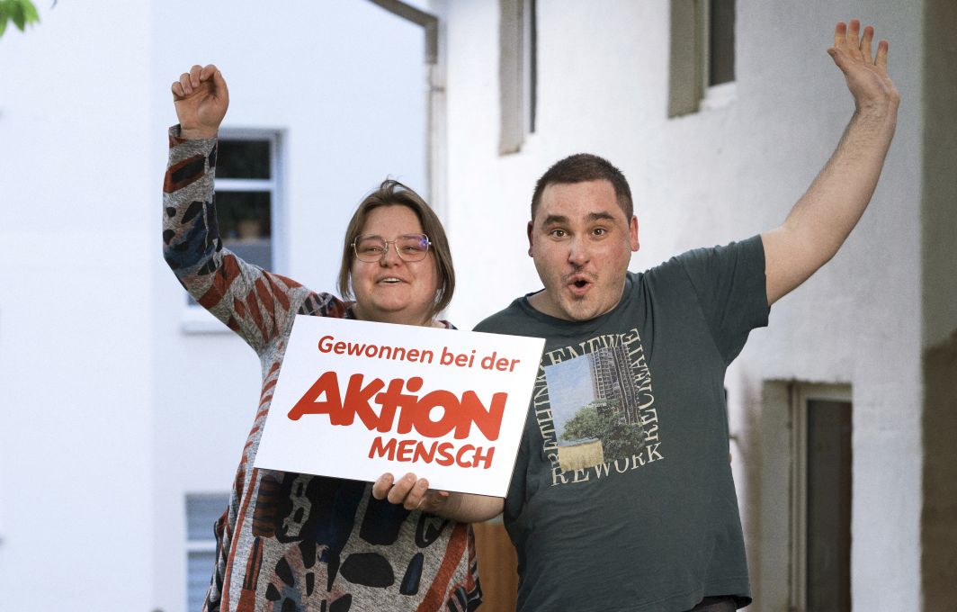 Felix und Luisa halten ein Schild zwischen sich mit der Aufschrift: Gewonnen bei der Aktion Mensch. Sie recken jeweils einen Arm jubelnd in die Luft.