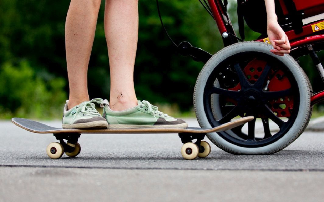 Zwei Füße stehen auf einem Skateboard, daneben fährt ein Rollstuhl.