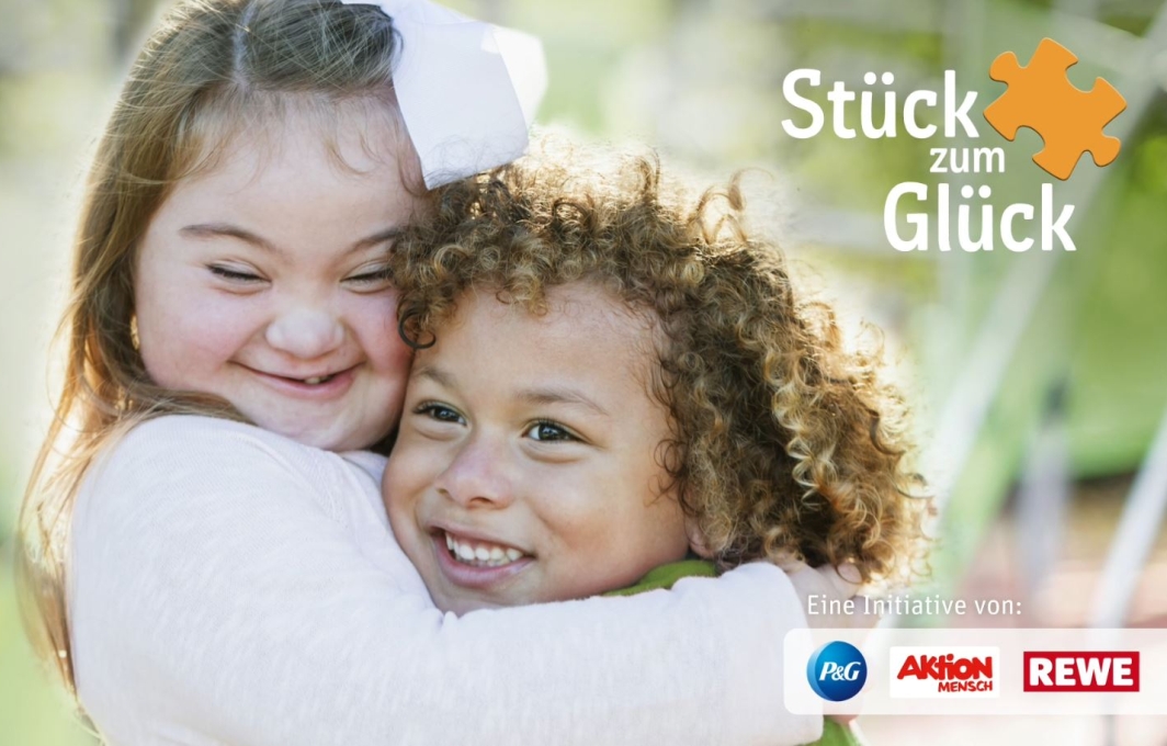 Ein Mädchen mit Down-Syndrom umarmt seine Freundin. Beide lachen. Daneben steht: Stück zum Glück. Eine Initiative von der Aktion Mensch, Procter & Gamble und Rewe.